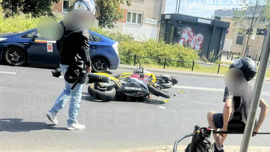 Motocykl leży na jezdni, a obok stoi mężczyzna w kurtce skórzanej, obserwujący sytuację. W tle widać zaparkowany samochód.