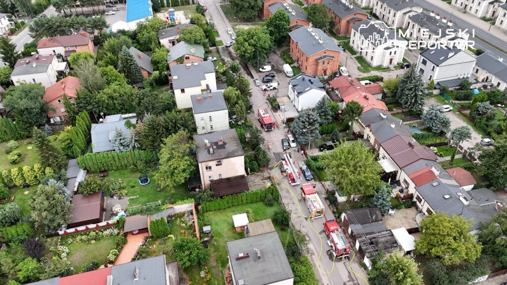 Wielu strażaków pracuje nad gaszeniem pożaru w zabudowanej dzielnicy, otoczonej zielenią i domami jednorodzinnymi.