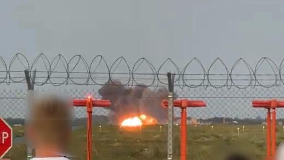 Osoby stojące za ogrodzeniem z drutu kolczastego obserwują wybuch na terenie lotniska.