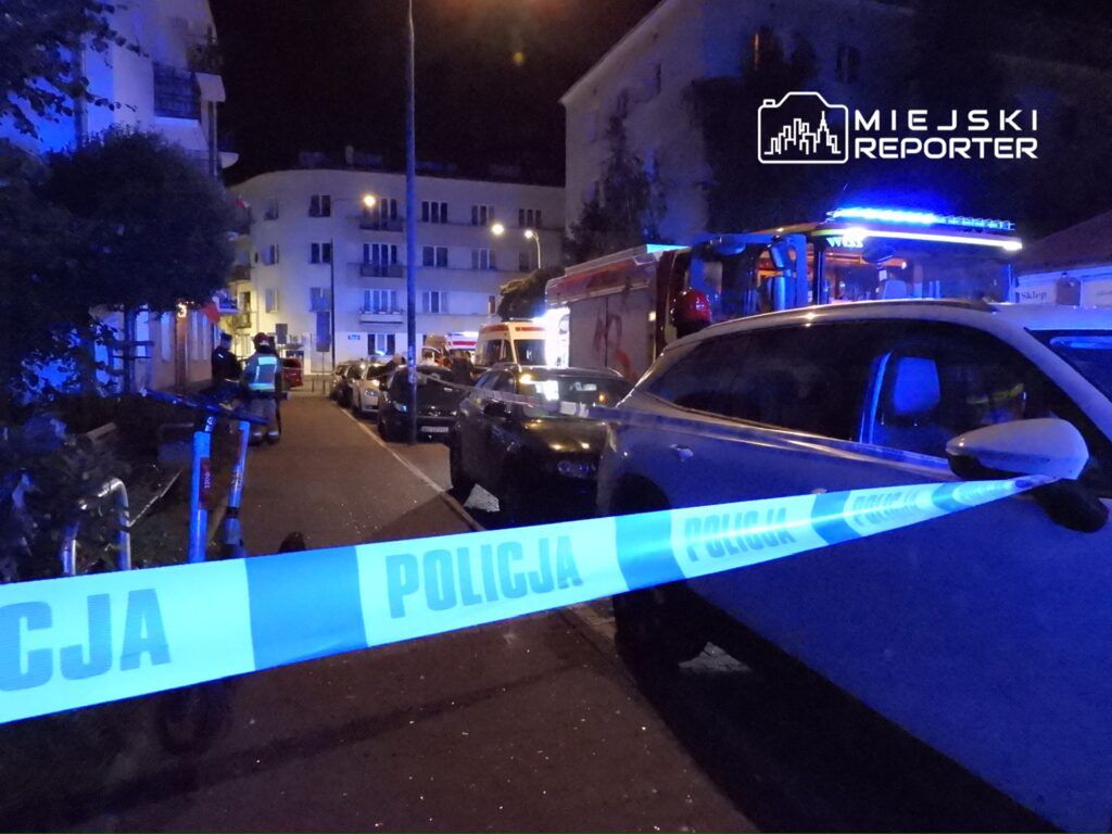 Policjanci zabezpieczają miejsce zdarzenia, a w tle widoczny wóz strażacki na oświetlonej ulicy nocą.