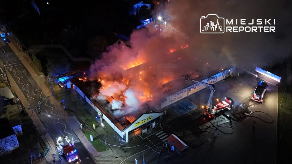 Płonący budynek sklepu Netto, otoczony przez strażaków i pojazdy ratunkowe, wypełniony gęstym dymem.