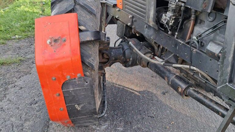 Traktor Ursus z uszkodzoną tylną oponą stoi na drodze, obok przewrócone auto osobowe oraz strażacy w mundurach.