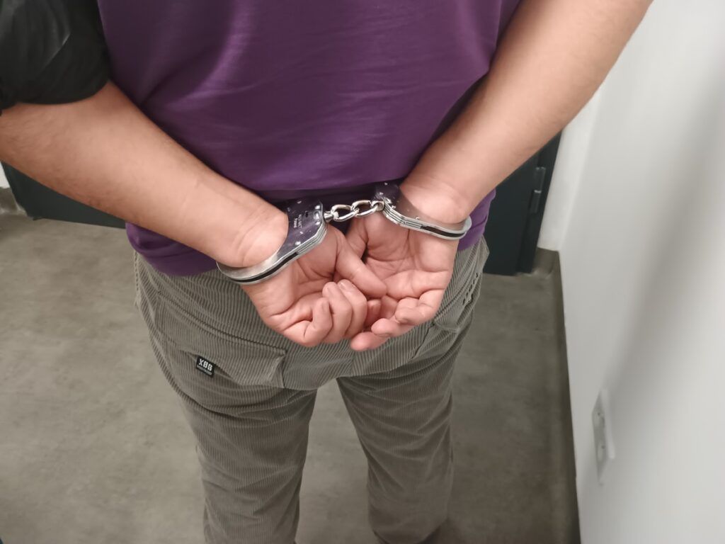 Mężczyzna w szarych spodniach ma założone kajdanki, stojąc w pomieszczeniu z betonową posadzką.