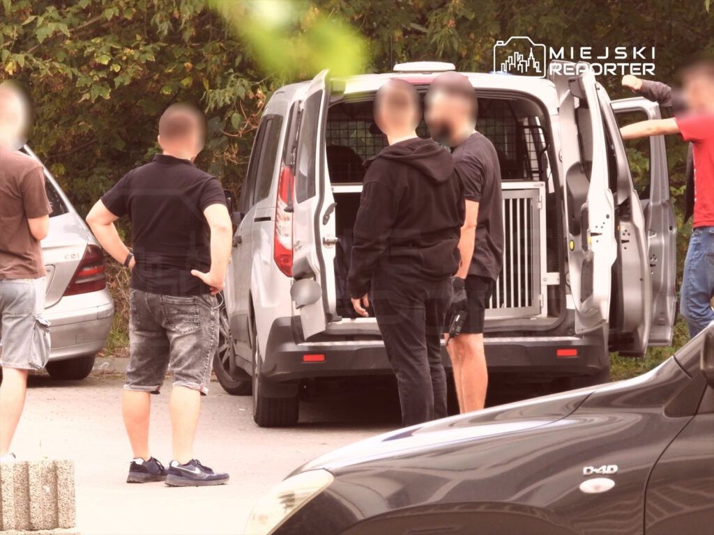 Grupa mężczyzn w różnych strojach rozmawia obok otwartego samochodu dostawczego z metalową klatką w środku.