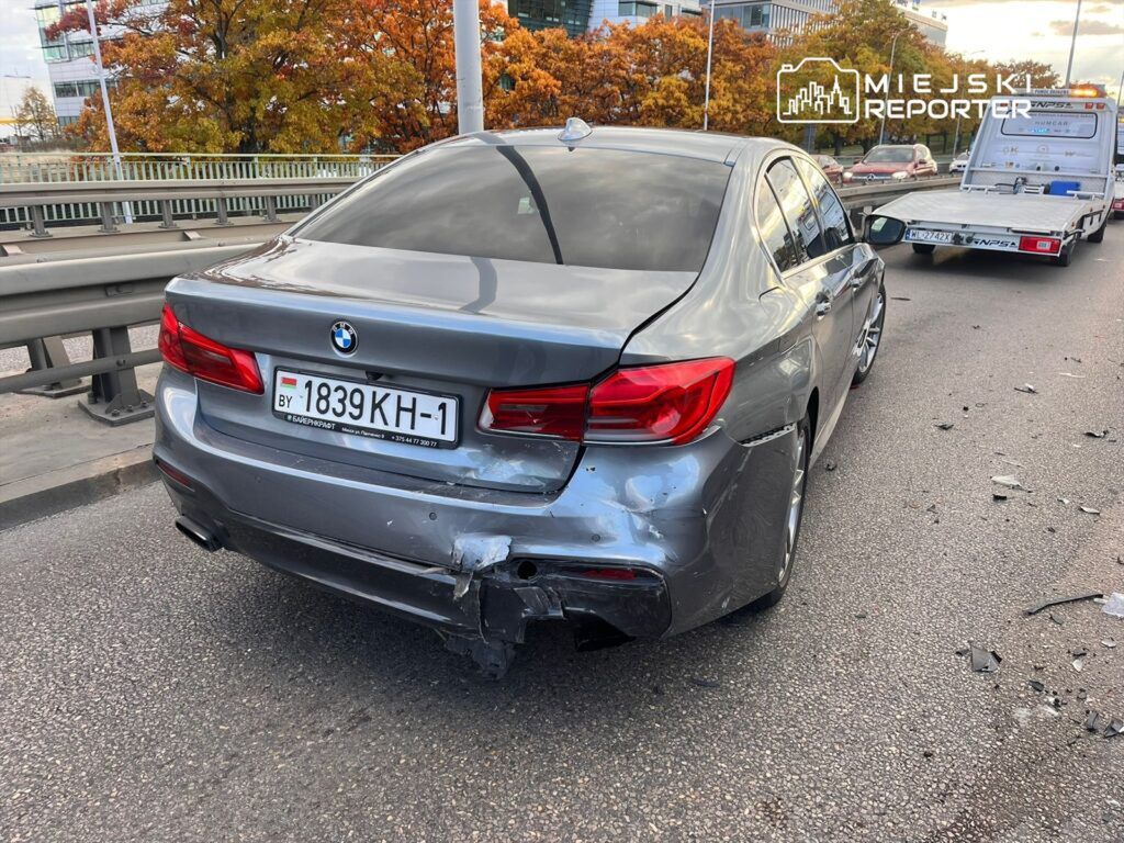 Uszkodzony samochód osobowy marki BMW z wgniecioną tylną częścią stoi na jezdni, obok przyczepa holownicza.