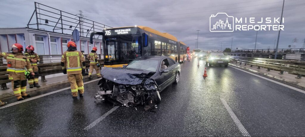 Grupa strażaków w mundurach z odblaskami ocenia skutki wypadku drogowego na mokrej jezdni, obok uszkodzonego samochodu osobowego.
