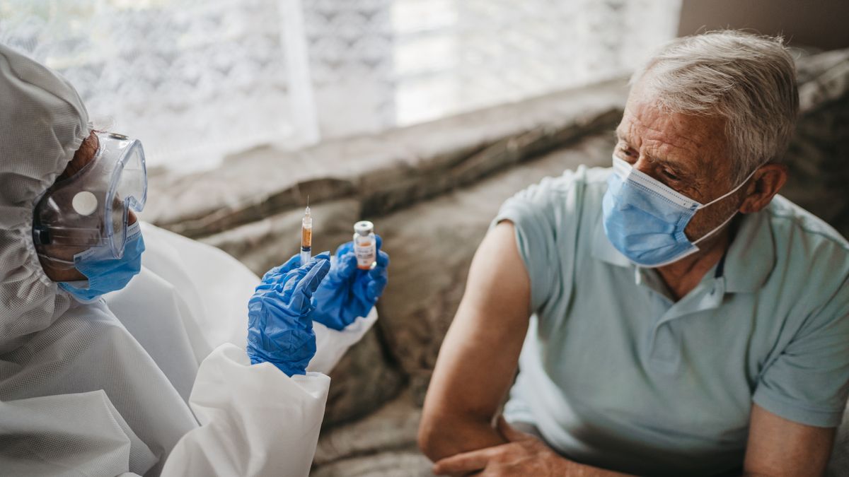 Female doctor in protective suit giving vaccine against covid19 virus to senior man at home