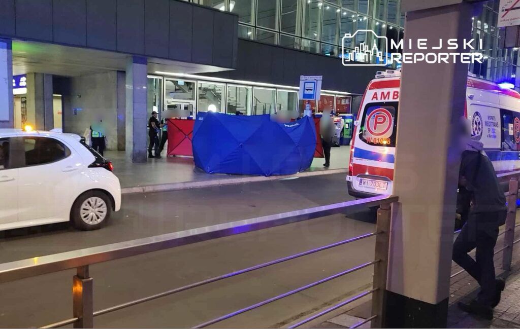 Policjanci zabezpieczają miejsce zdarzenia przy nowoczesnym budynku, obok stojący ambulans i biały samochód osobowy.