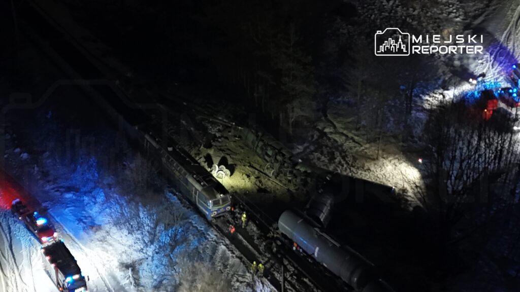 Pojazdy ratunkowe z niebieskimi światłami pracują przy wykolejonym pociągu na zaśnieżonej linii kolejowej.