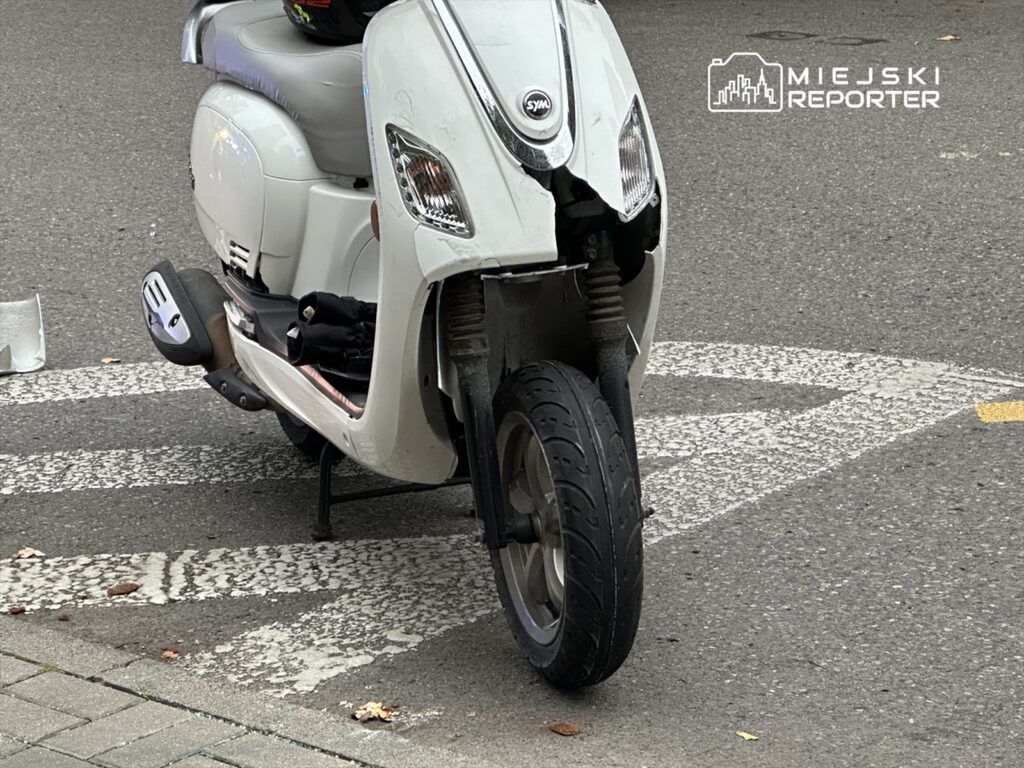 Uszkodzony skuter zaparkowany na przejściu dla pieszych, z widoczną uszkodzoną przednią częścią.