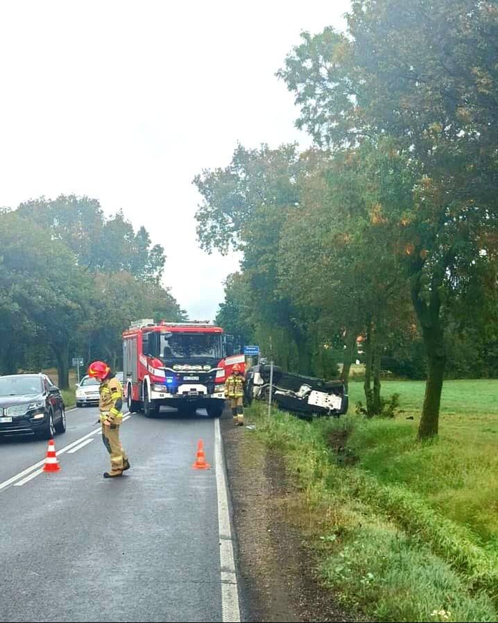 Strażacy pracują przy przewróconym pojeździe obok drogi, podczas gdy inne samochody przejeżdżają obok.