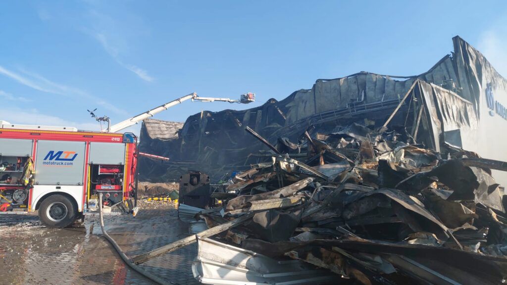 W tle zniszczony budynek po pożarze, z przodu wóz strażacki z rozwiniętym wężem, gaszący pozostałości ognia.