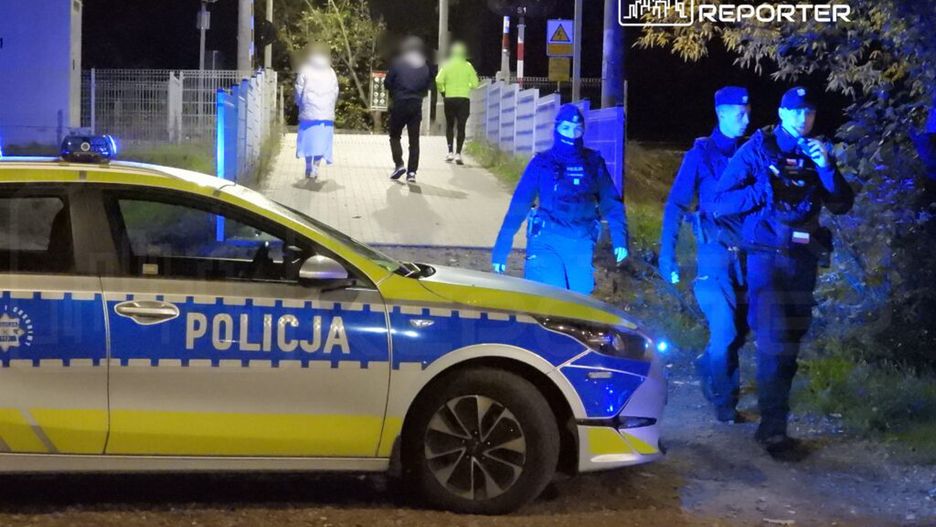 Policjanci w mundurach patrolują okolicę nocą, obok radiowozu z włączonymi światłami, w tle piesi przechodzą przez most.