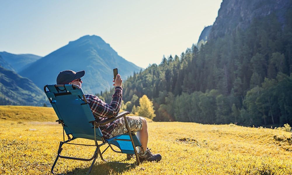 Mężczyzna siedzący na krześle campingowym, trzymający telefon w rękach, z widokiem na górski krajobraz.