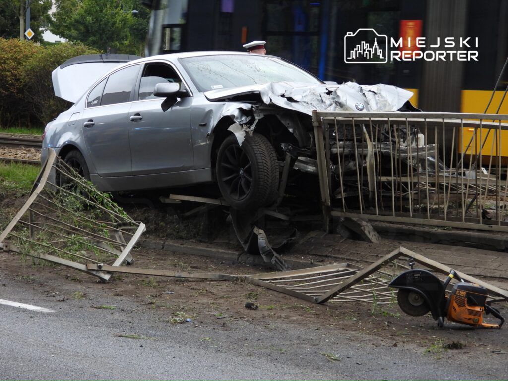 Uszkodzony srebrny samochód osobowy stoi na krawędzi drogi, z uszkodzoną przednią częścią i zniszczonym ogrodzeniem. W tle widać tramwaj.