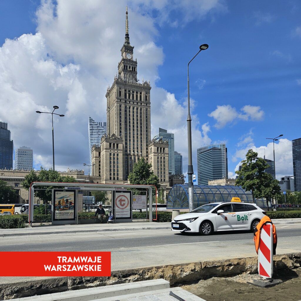 Biały samochód marki Bolt przejeżdża obok przystanku tramwajowego z nowoczesnym biurowcem w tle.