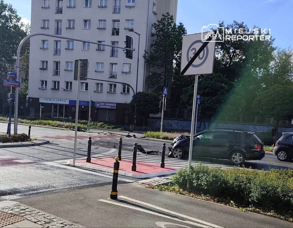 Zniszczony samochód na skrzyżowaniu, obok znak drogowy z ograniczeniem prędkości i budynek mieszkalny w tle.