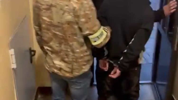 Mężczyzna w mundurze trzyma rękę skuta w kajdankach młodego mężczyzny, wychodząc z budynku.