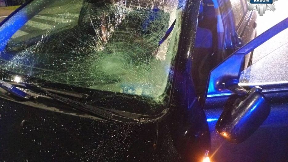 Uszkodzony samochód z rozbitą szybą przednią, zaparkowany w deszczowym otoczeniu nocą. W tle widać niebieskie światła policyjne.