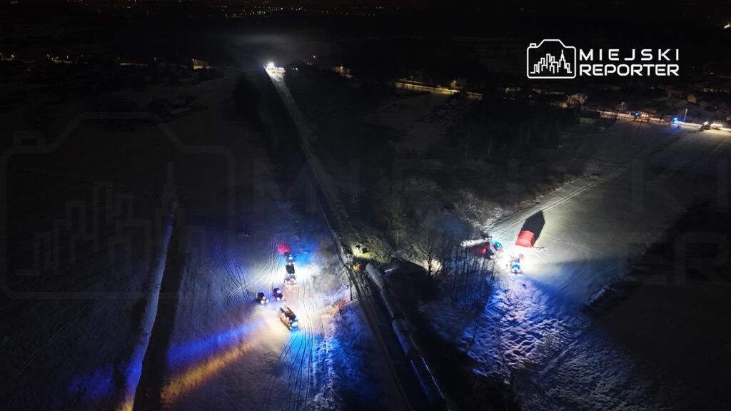 Grupa ratunkowa pracuje przy przewróconym wagonie kolejowym na ośnieżonym terenie w nocy, używając niebieskich i czerwonych świateł.