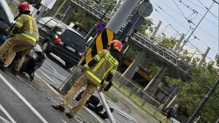 Dwóch strażaków w mundurach zajmuje się usuwaniem skutków wypadku drogowego przy zniszczonym słupie.