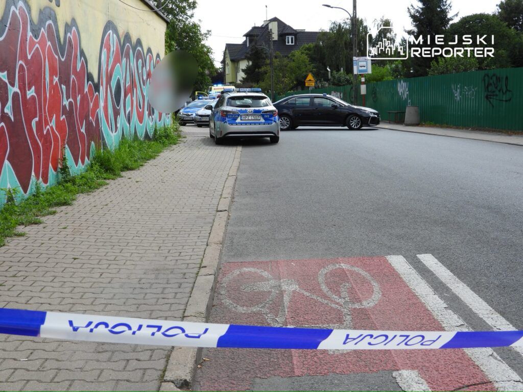 Policyjny radiowóz zaparkowany obok graffiti na murze, z taśmą zabezpieczającą wzdłuż chodnika.
