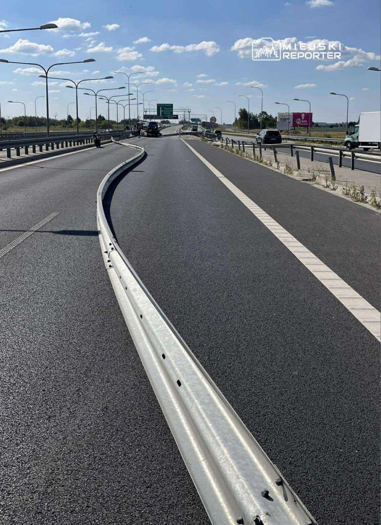 Zakrzywiona bariera ochronna na szerokiej, asfaltowej jezdni, w tle oznakowanie drogi i pojazdy.