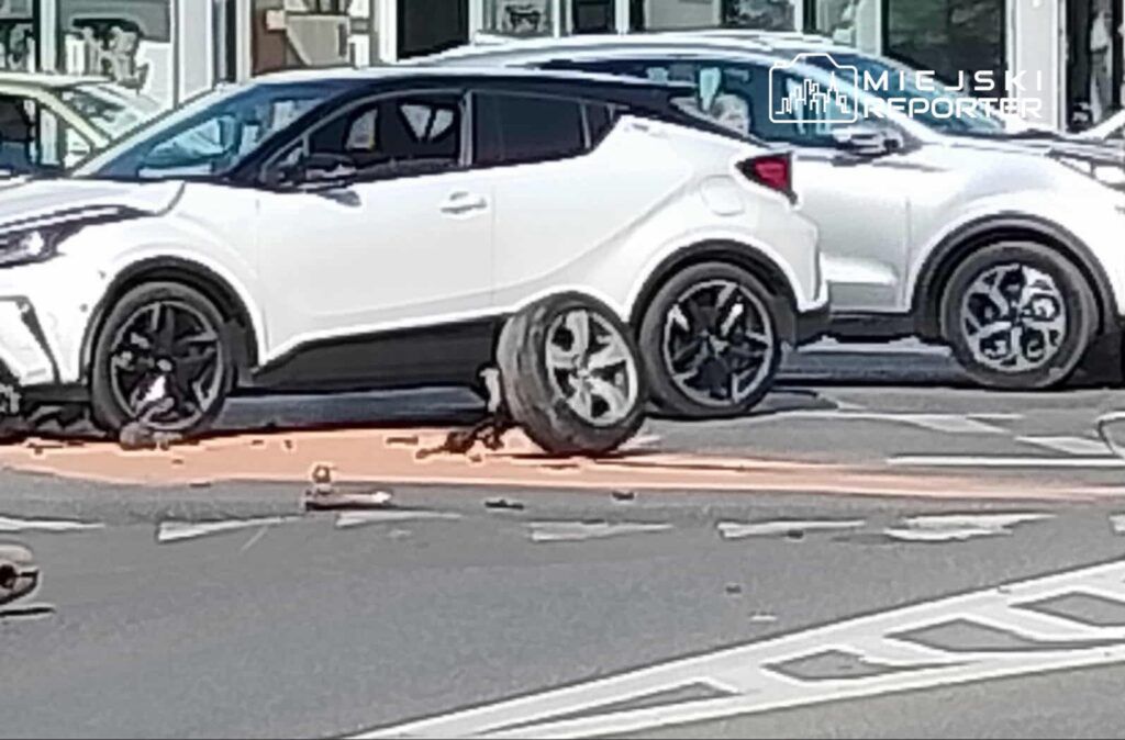 Biały samochód osobowy z uszkodzonym kołem stoi na drodze, obok inny pojazd. Na jezdni widać resztki części.