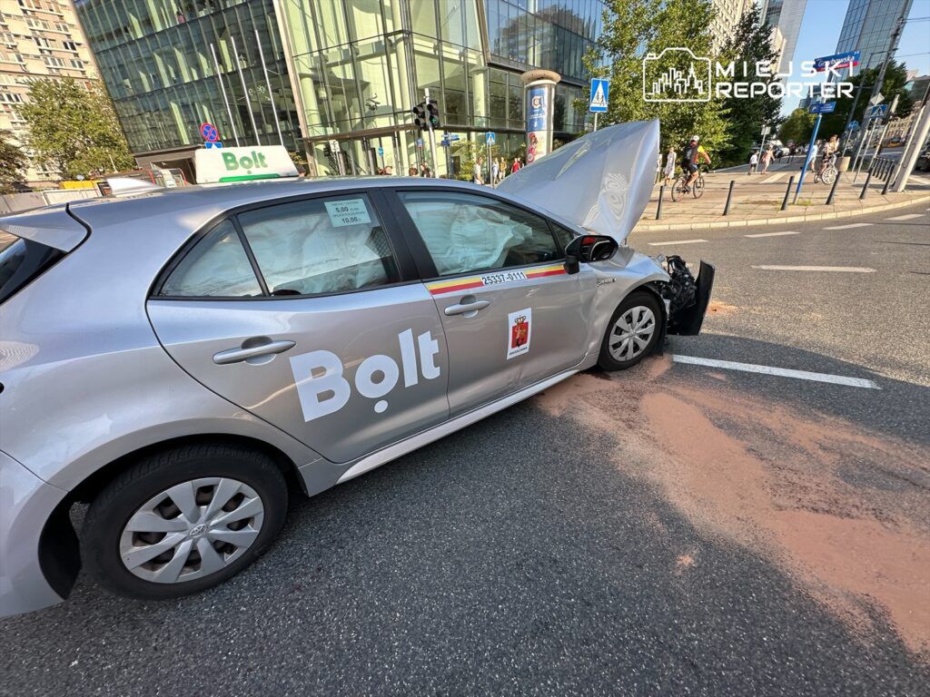 Samochód marki Bolt z uszkodzoną maską stoi na ulicy, obok nowoczesnego biurowca, na asfalcie widoczny proszek.