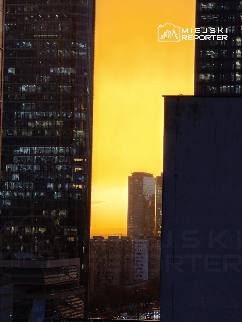 Złota poświata zachodzącego słońca między nowoczesnymi wieżowcami w miejskiej scenerii.