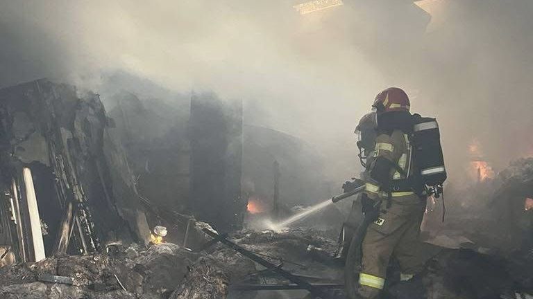 Strażak w pełnym ekwipunku gaśniczym ugasza pożar w zadymionym pomieszczeniu z zniszczonymi meblami.