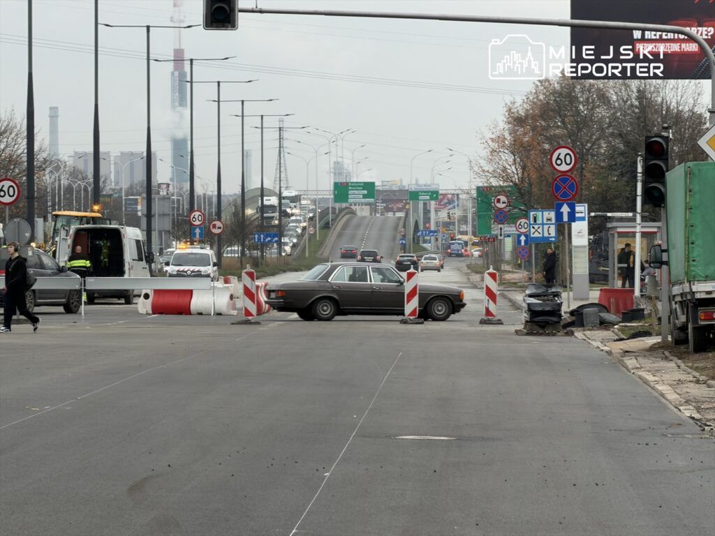 Samochód osobowy zablokowany na drodze, obok prace drogowe i oznakowanie. W tle widoczny ruch uliczny.