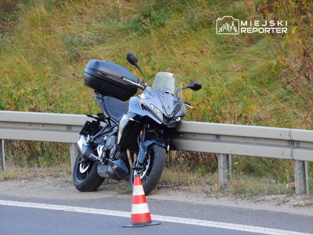 Czarny motocykl zaparkowany przy barierce ochronnej, oznaczony trójkątnym znakiem ostrzegawczym.