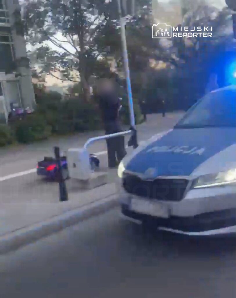 Policjant w mundurze stoi obok radiowozu na ulicy, a w tle widać zieleń i budynki.