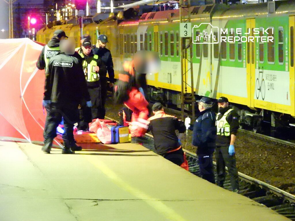 Ratownicy medyczni udzielają pomocy osobie leżącej na peronie przy torach kolejowych, w nocy.