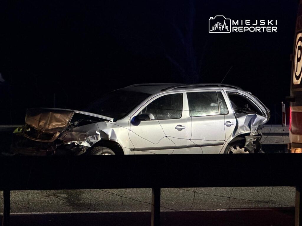 Uszkodzony samochód osobowy z wgniecionym przodem i tyłem stoi na drodze w nocy, obok barierki.