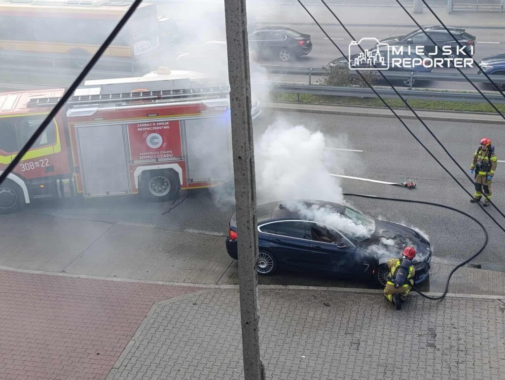 Strażacy gaszą palący się samochód osobowy na poboczu drogi, w tle widoczna strażacka ciężarówka.
