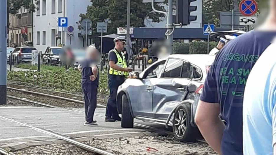 Policjant rozmawia z kierowcą uszkodzonego samochodu na skrzyżowaniu obok torów tramwajowych.