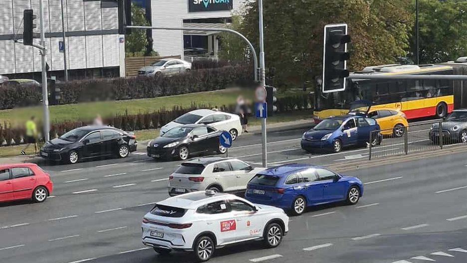 Ruchliwa ulica z kilkoma samochodami, w tym białym SUV-em, oraz przystanek autobusowy z żółtym autobusem.