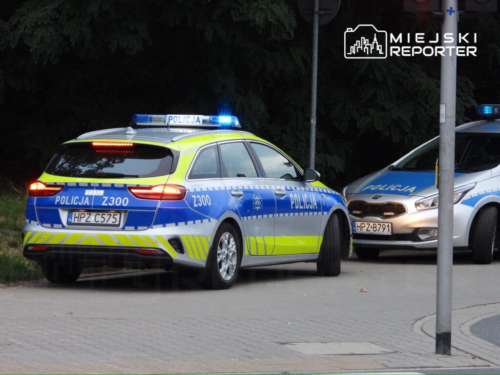 Dwa radiowozy policyjne zaparkowane na chodniku, z włączonymi sygnałami świetlnymi w otoczeniu drzew.