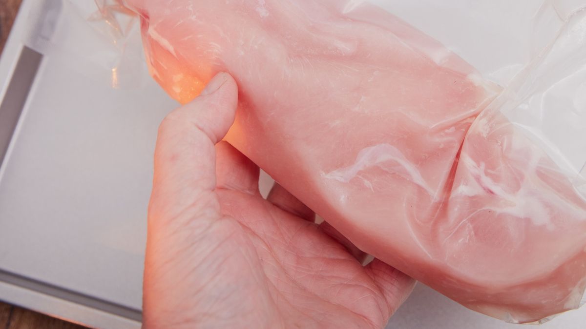 Vacuum-sealed chicken filletsHuman hand holding three in polyethylene bag vacuum-sealed chicken fillets. Jelly roll pan on teak wood surface. High point of view.annick vanderschelden photograph