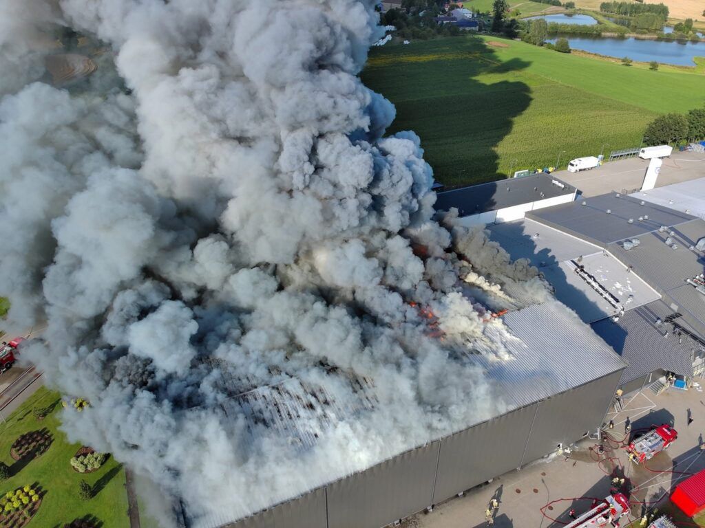 Gęsty dym wydobywający się z dachu dużego budynku przemysłowego, w pobliżu widać wozy strażackie.