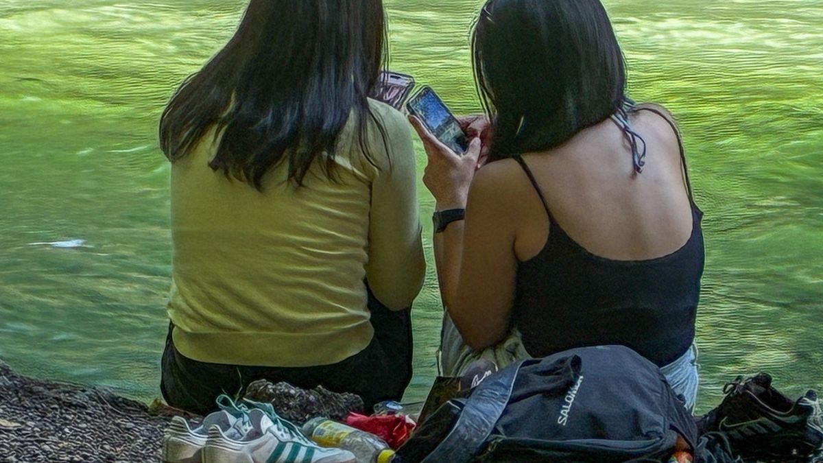 Two young women sit by a stream shore using their smartphones in a park in Munich, Bavaria, Germany, on August 11, 2025. (Photo by Michael Nguyen/NurPhoto via Getty Images)