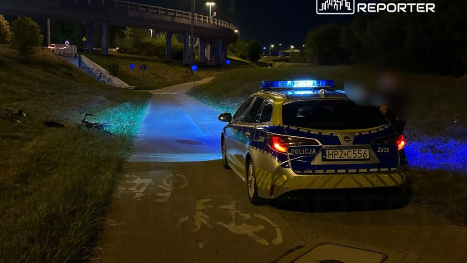 Policyjny radiowóz z włączonymi sygnalizatorami zatrzymany na ścieżce rowerowej w nocy. W tle widoczna droga i most.
