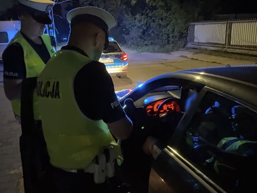 Dwaj policjanci w odblaskowych kamizelkach kontrolują kierowcę w samochodzie nocą. W tle widać radiowóz.