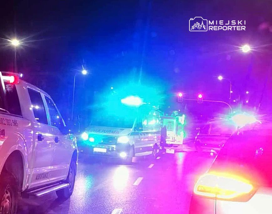 Nocna scena na ulicy z pojazdami służb ratunkowych, w tym karetka i samochód strażacki, z włączonymi sygnałami świetlnymi.