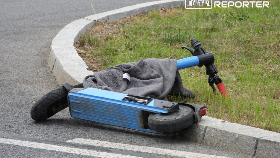 Złamana hulajnoga leży na poboczu drogi, przykryta szarym ręcznikiem, obok trójkąt ostrzegawczy.