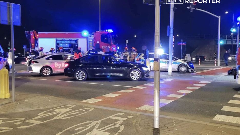 Na skrzyżowaniu z sygnalizacją świetlną stoi czarny samochód, wokół niego zebrani policjanci i strażacy. W tle widoczny wóz strażacki z włączonymi sygnałami świetlnymi.