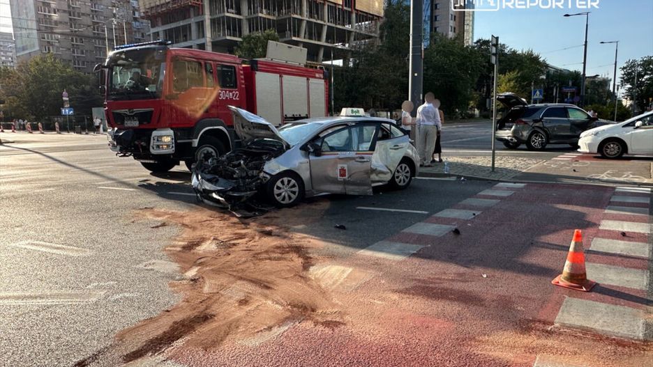 Zniszczony samochód osobowy stoi na skrzyżowaniu, obok wozu straży pożarnej, a kierowcy rozmawiają na chodniku.