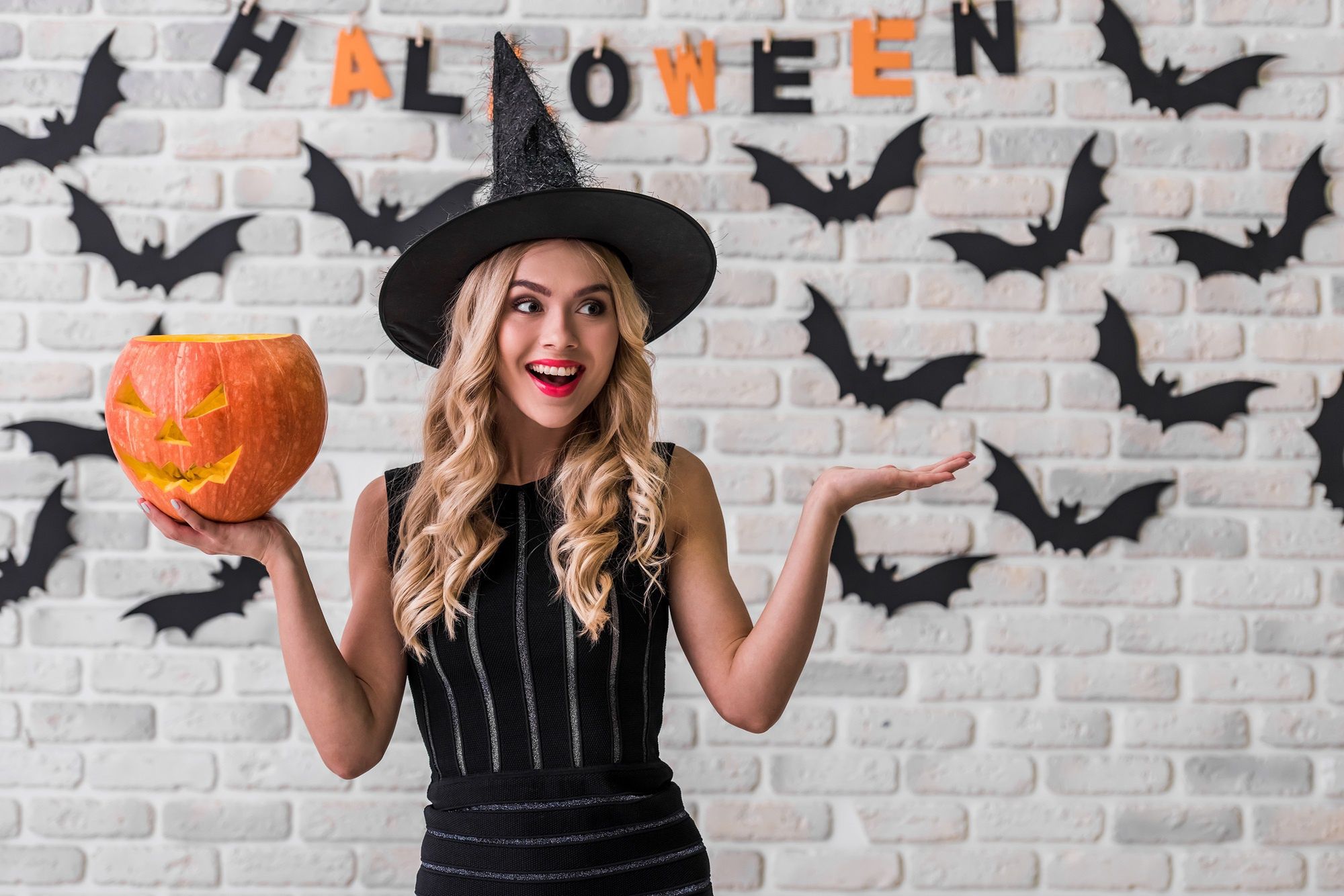 Beautiful blonde girl in black dress and witch hat is holding a scary pumpkin, looking away and smiling, on background decorated for Halloweenadult, attractive, background, bat, beautiful, beauty, black, blonde, carnival, caucasian, celebration, costume, dark, decoration, demon, dress, expression, fashion, female, fun, girl, glamour, halloween, happy, hat, hold, holiday, jack o'lantern, lips, model, party, people, playful, portrait, posing, pretty, pumpkin, red, scary, smile, studio, style, stylish, white, witch, woman, young, adult, attractive, background, bat, beautiful, beauty, black, blonde, carnival, caucasian, celebration, costume, dark, decoration, demon, dress, expression, fashion, female, fun, girl, glamour, halloween, happy, hat, hold, holiday, jack o'lantern, lips, model, party, people, playful, portrait, posing, pretty, pumpkin, red, scary, smile, studio, style, stylish, white, witch, woman, young